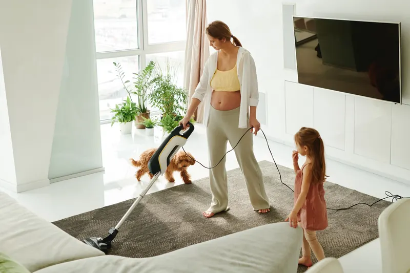 Pregnant mother cleaning carpet with vacuum as daughter and dog play indoors
